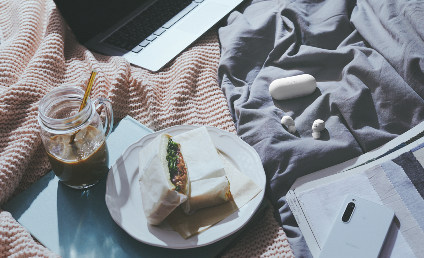 Imagen de los audífonos WF-C700N de color blanco y su estuche sobre una manta de pícnic con comida, bebida, una laptop y un teléfono